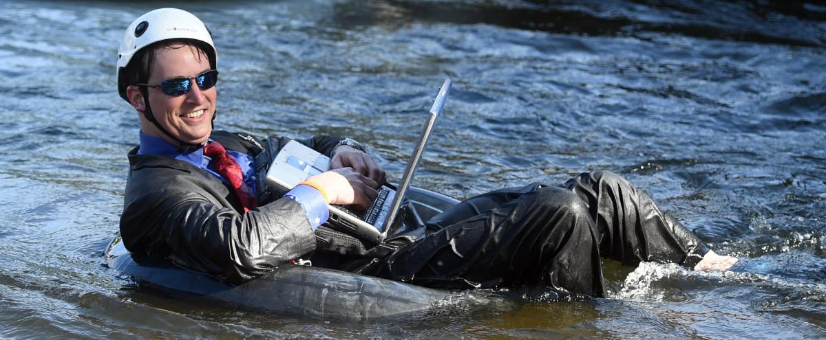 Man wearing a white helmet in a suit holding a laptop floating down the river